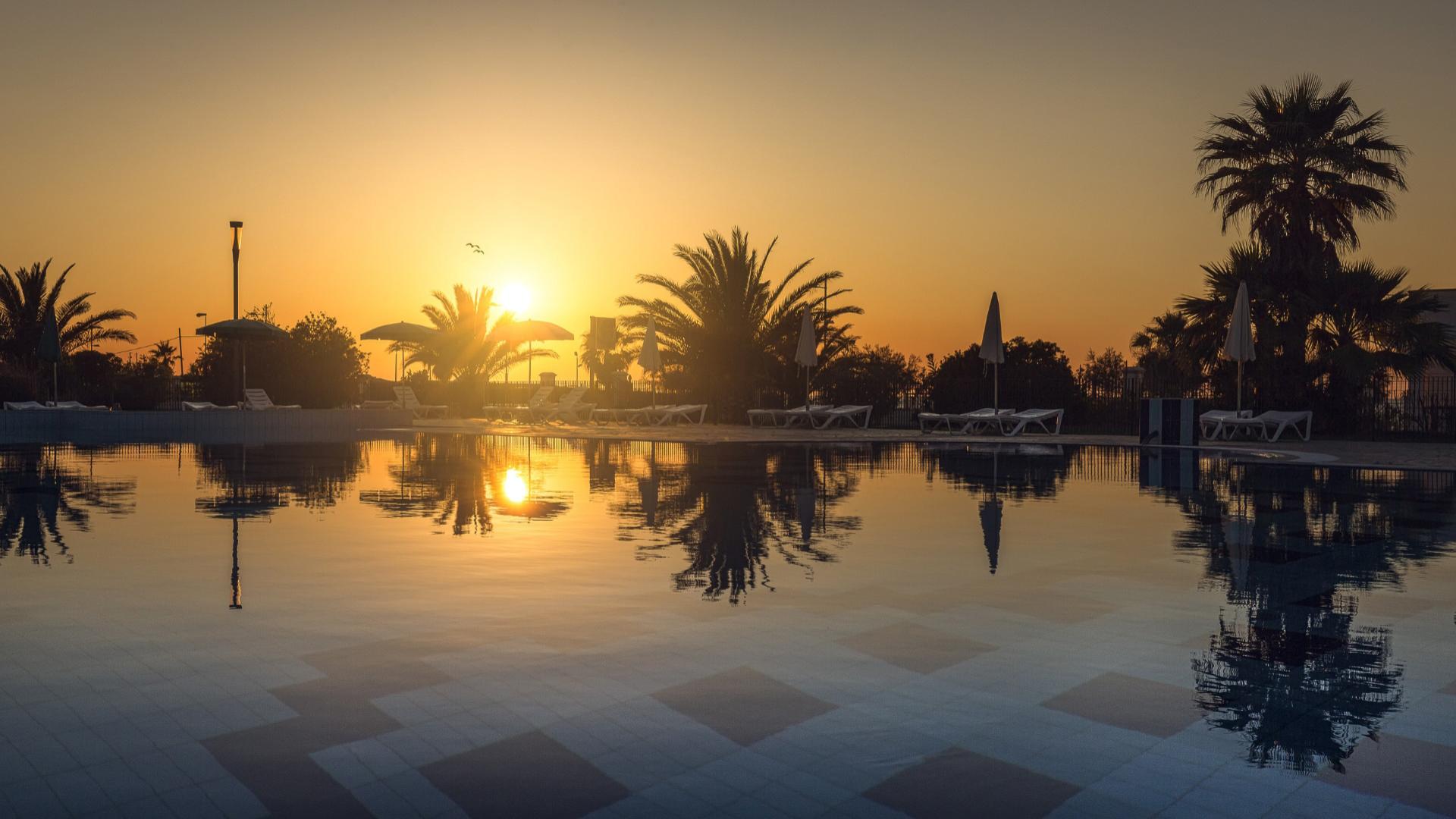 Tramonto su piscina con palme e lettini riflessi nell'acqua.