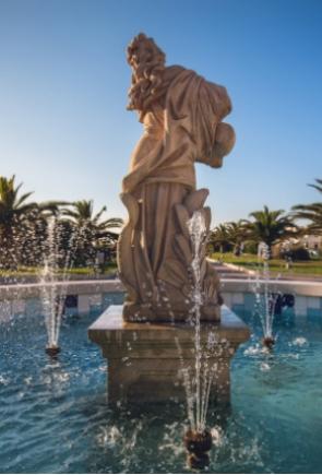 Statua in una fontana con spruzzi d'acqua, circondata da palme.