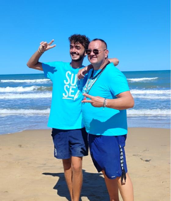 Due persone sorridenti in spiaggia, vestite di blu, fanno il segno di pace.