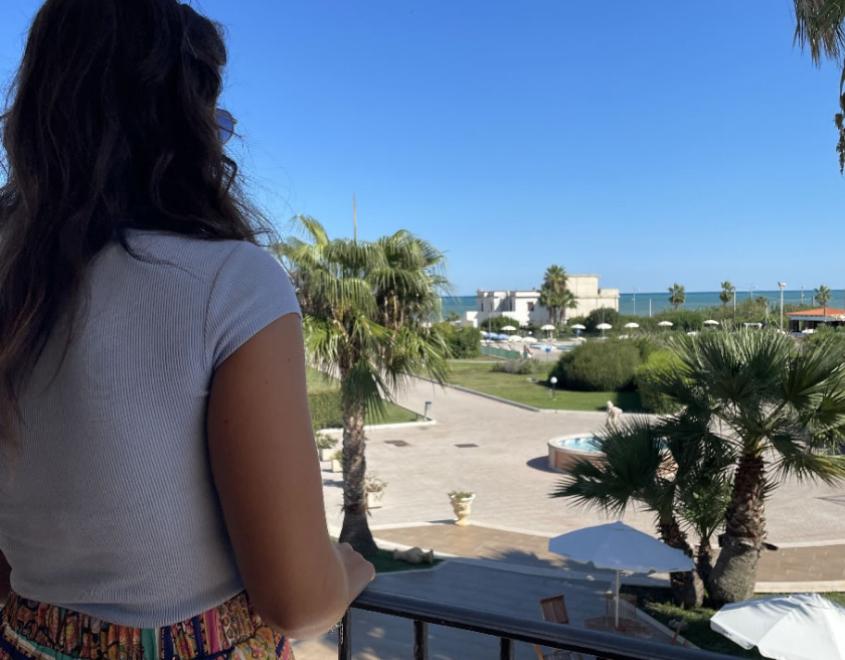 Donna osserva il mare da un balcone, circondata da palme e cielo azzurro.