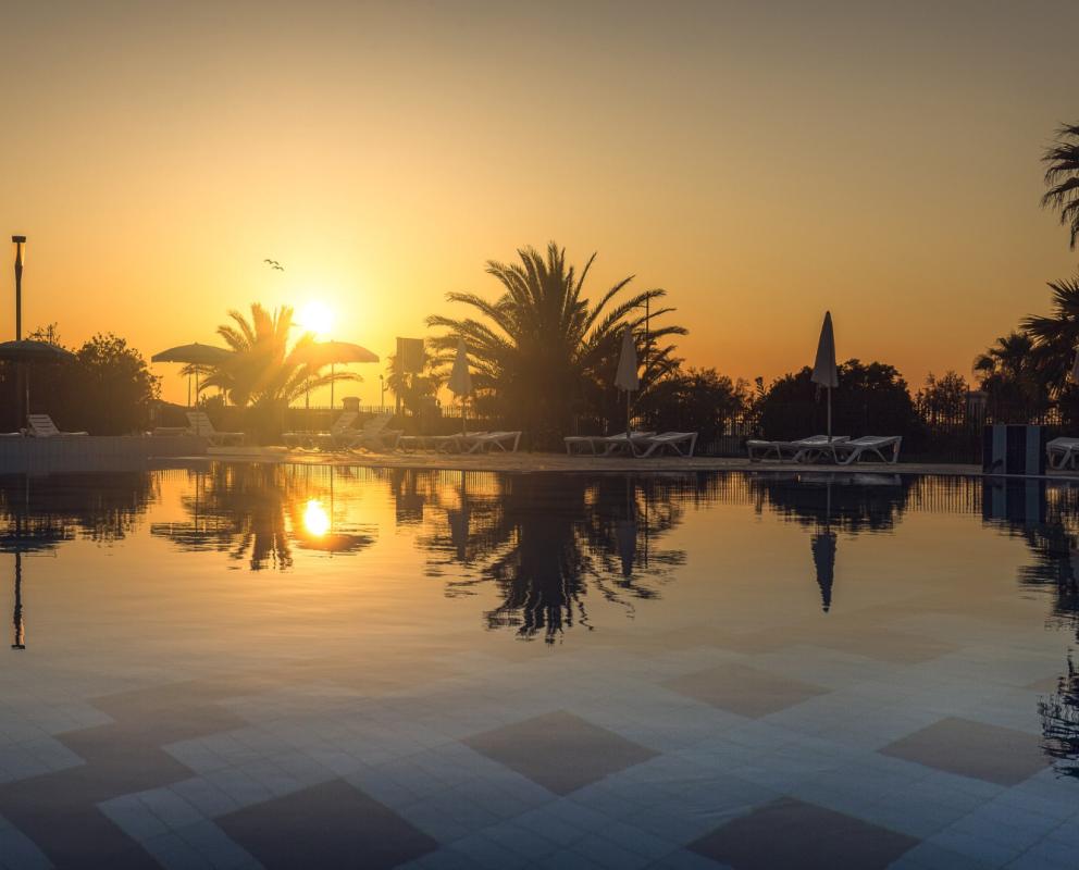 Tramonto su piscina con palme e lettini riflessi nell'acqua.